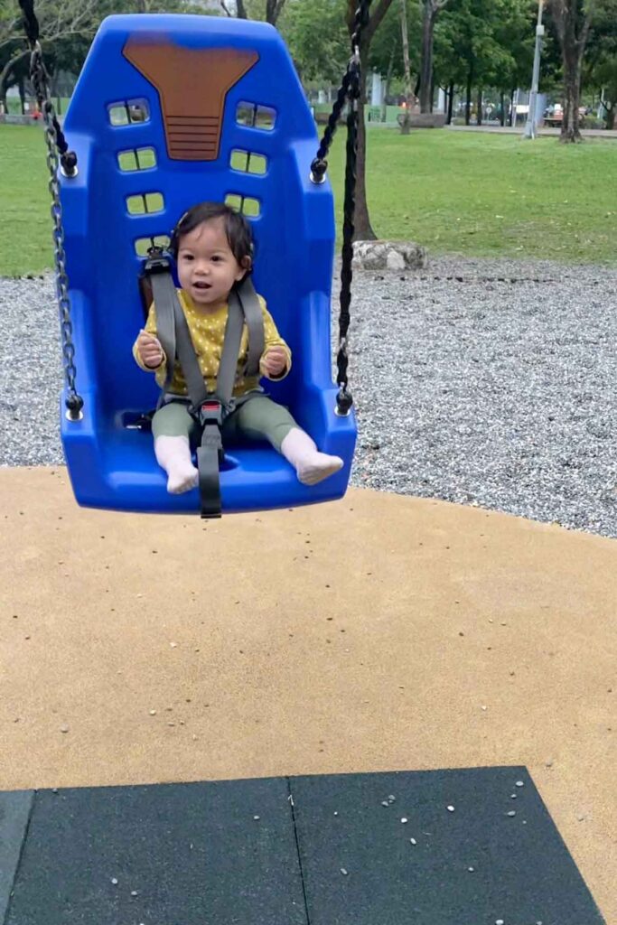 A smiling toddler is secured in a blue adaptive swing with safety harnesses at a playground, surrounded by rubber and gravel surfacing, with grass and trees in the background.