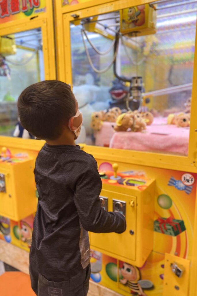 A young boy wearing a mask and dark long sleeve shirt stands in front of a bright yellow claw machine, focused on the plush toys inside as the claw lowers toward a pile of tan teddy bears.