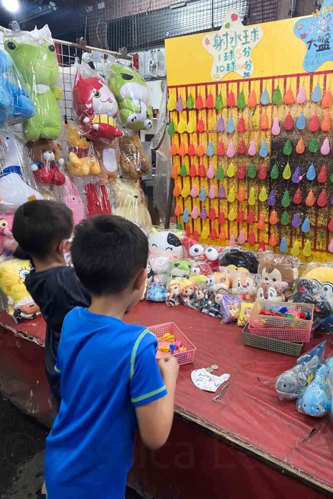 Two boys play a dart balloon game at a night market stall, aiming at a wall of colorful water balloons, with shelves of plastic-wrapped stuffed animals and toys displayed around it.