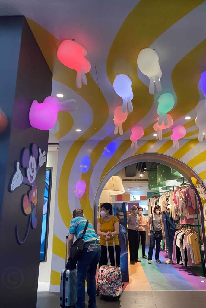 Shoppers walk through a whimsically decorated children’s clothing store with a curvy striped yellow and white ceiling and colorful bunny-shaped ceiling lights. A large Mickey Mouse graphic is on the left wall, and some patrons are browsing with suitcases.