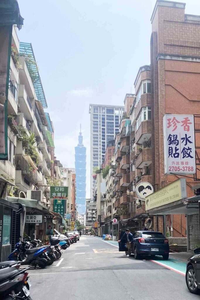 A narrow urban street in Taipei's Da-An district with scooters parked along the sides and mid-rise buildings flanking the road; Taipei 101 rises prominently in the distant background under a clear sky.