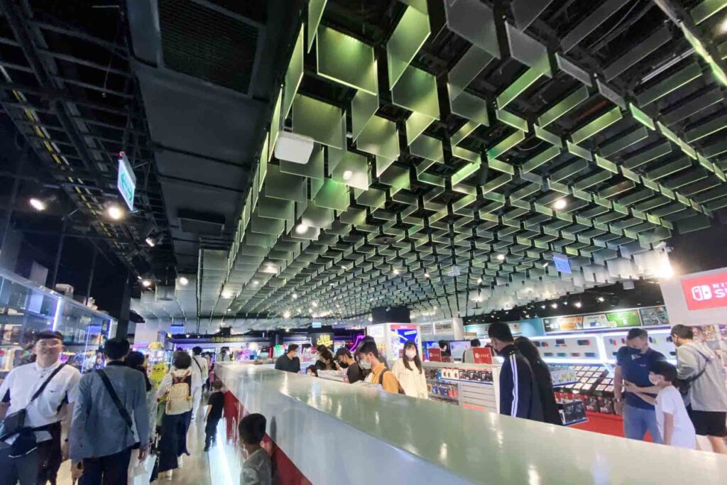 Wide, modern indoor space with a futuristic green-lit ceiling and rows of electronics booths; shoppers browse among gadgets, accessories, and Nintendo Switch products in Taipei’s electronics market.