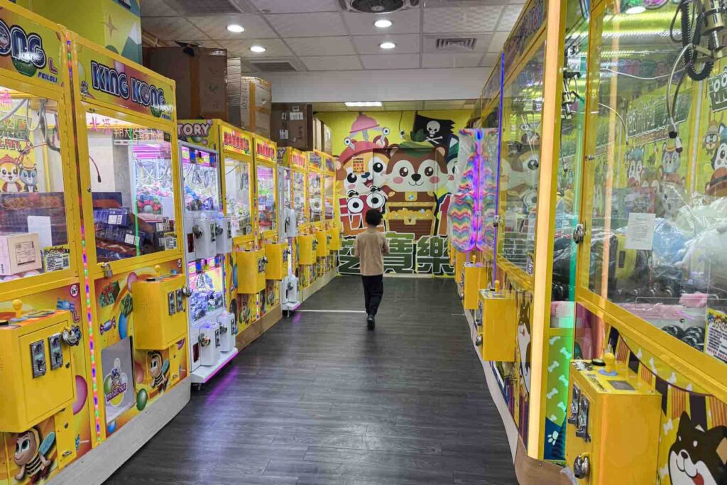 A young boy walks through an arcade filled with bright yellow claw machines in Taipei, each packed with prizes. A colorful cartoon mural of panda characters decorates the back wall.