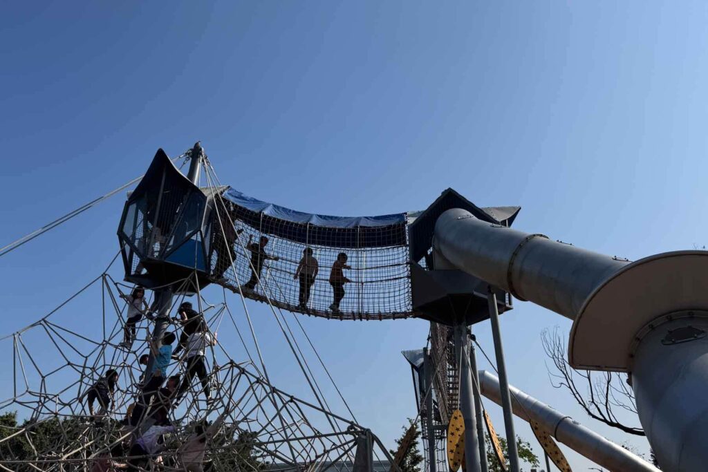 Children climb a large metal geometric structure and a crisscrossed and suspended bridge connected to tall towers with metal tube slides, all under a clear blue sky at Huabo Park in Taipei.