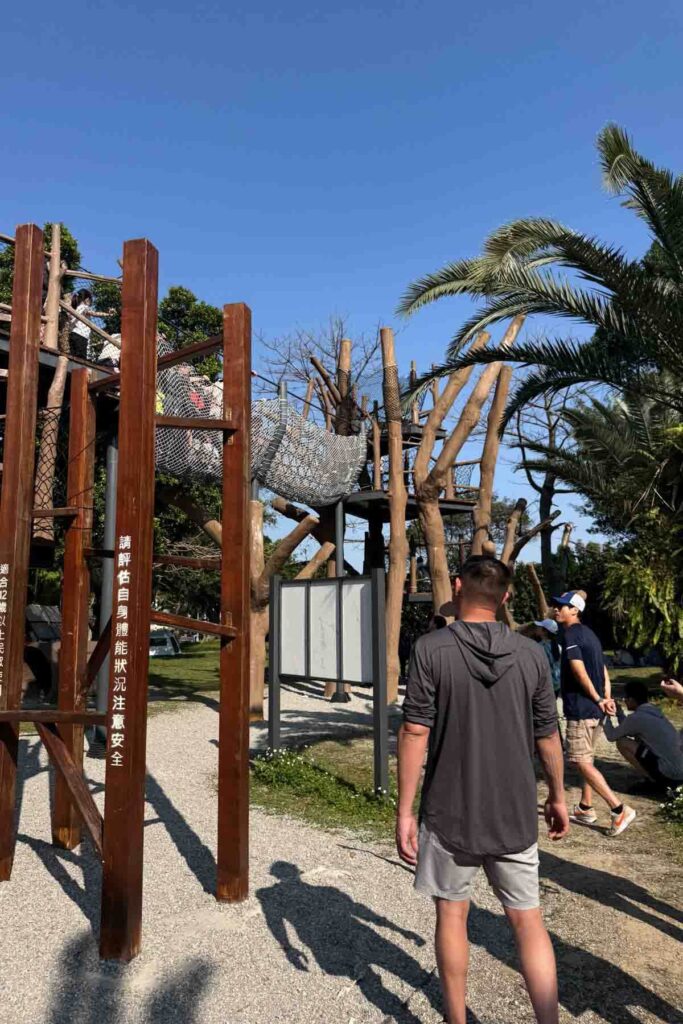 Adults and children gather around a large wooden playground structure in Taipei with rope bridges and nets nestled among palm trees and green space on a sunny day.