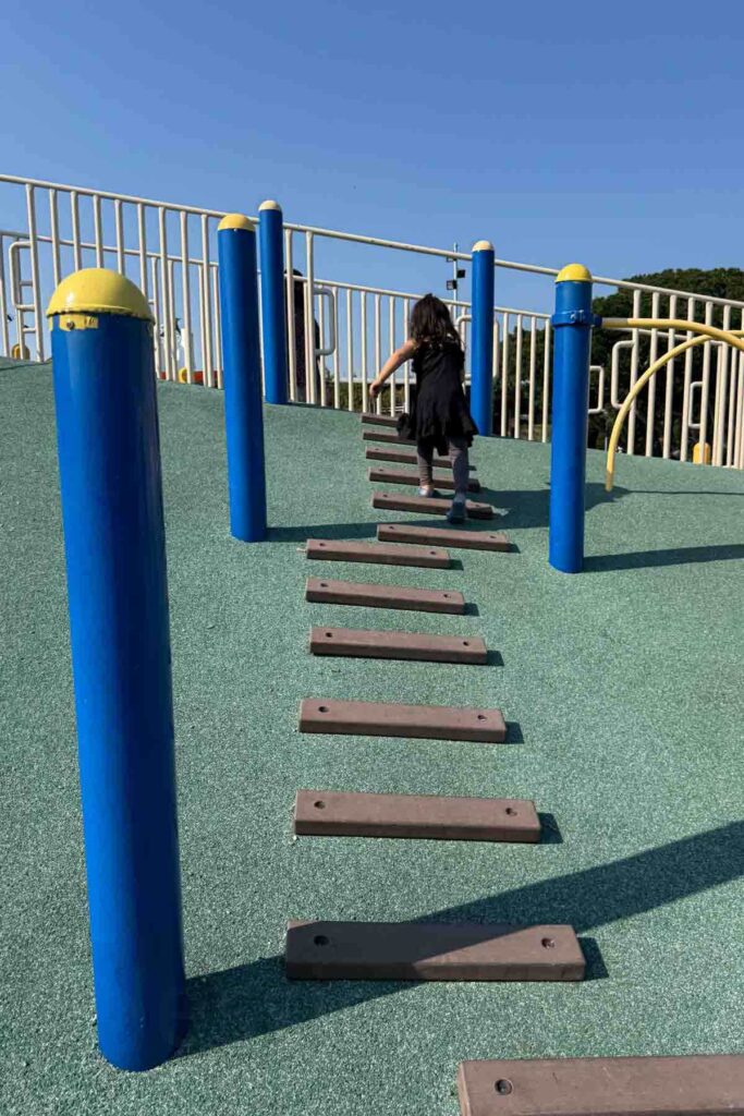 A young child in a black outfit climbs a steep, soft-surface playground hill using evenly spaced wooden steps, surrounded by blue and yellow posts under a bright blue sky at a Taipei playground.
