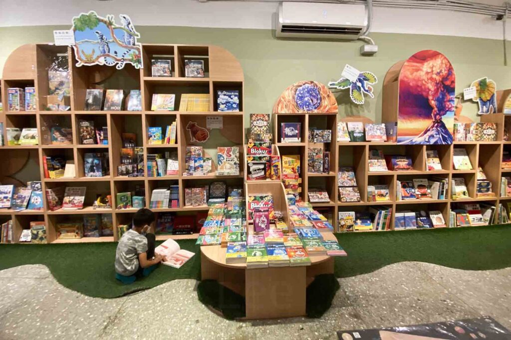 Colorful, organized wooden shelves packed with books and board games, with a child sitting on green turf reading a book; wall art includes volcano and animal illustrations at a bookstore in Huashan Park, Taipei.