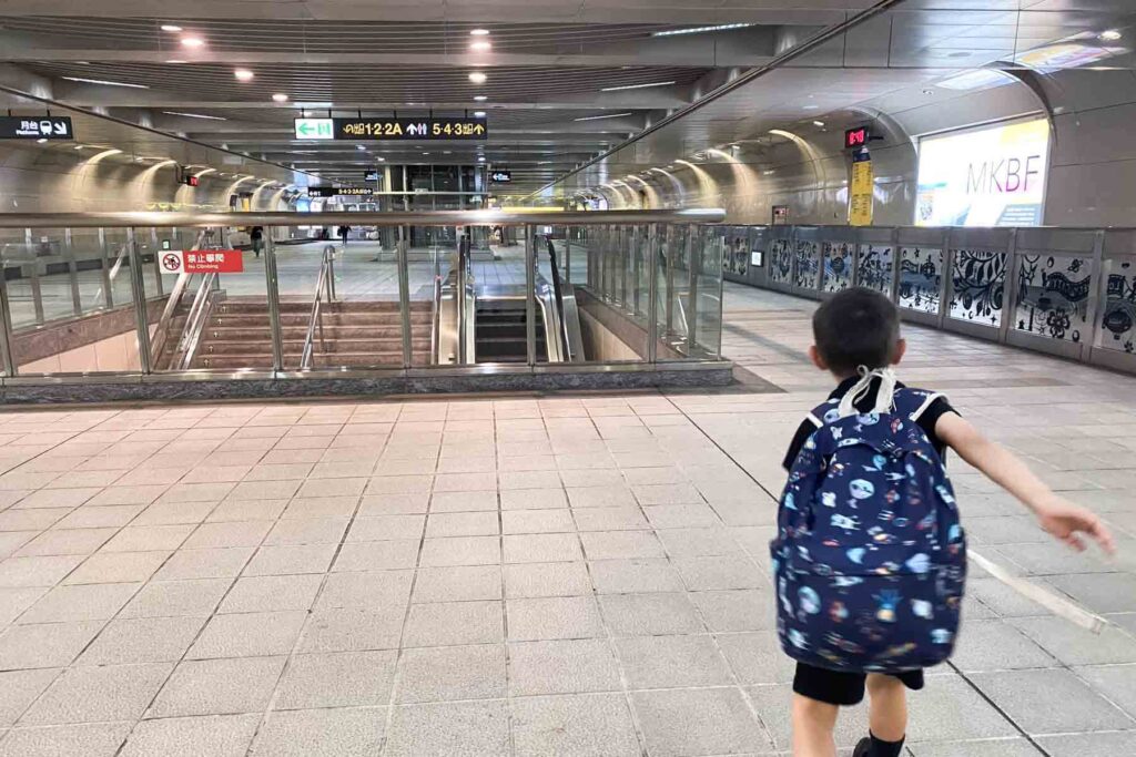 A young boy wearing a navy blue backpack dashes through an empty, modern Taipei metro station toward a set of escalators and stairways. Overhead signs display route numbers in Chinese and English.