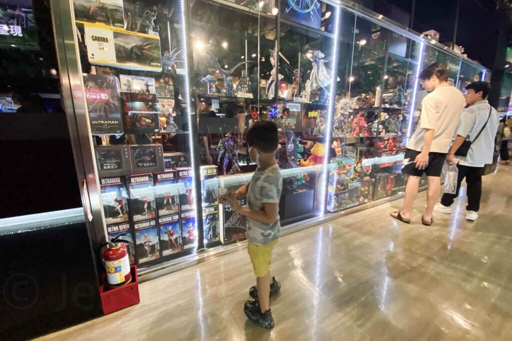 A boy in a gray camouflage shirt and yellow shorts stands in front of a lit-up display case packed with Ultraman action figures and collectibles in a Taipei store.