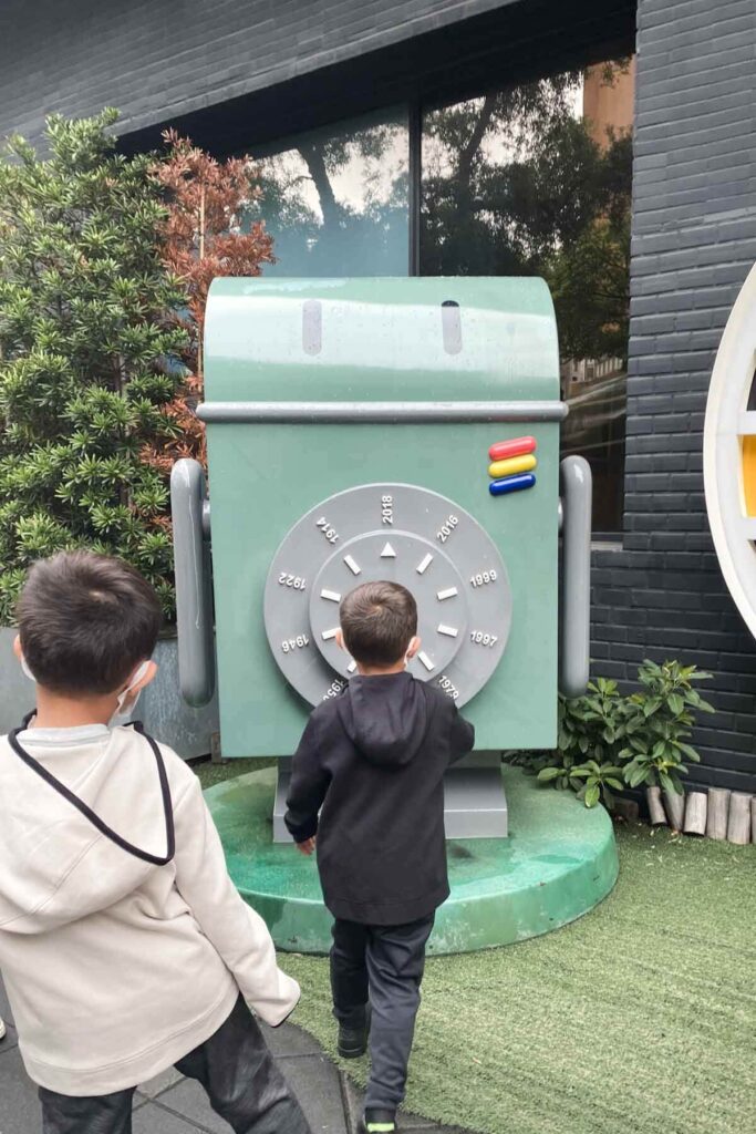 Two young boys explore a large green robot statue installation featuring a dial marked with years ranging from 1946 to 2016, resembling a time machine display, outside a modern building in Taipei.