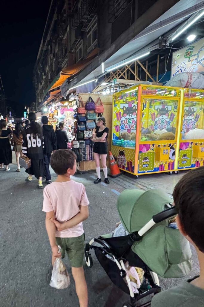 A bustling Taipei night market scene with a group of kids near a colorful "Lucky Catch" claw machine. Stalls selling handbags and accessories are lit brightly against the dark sky.