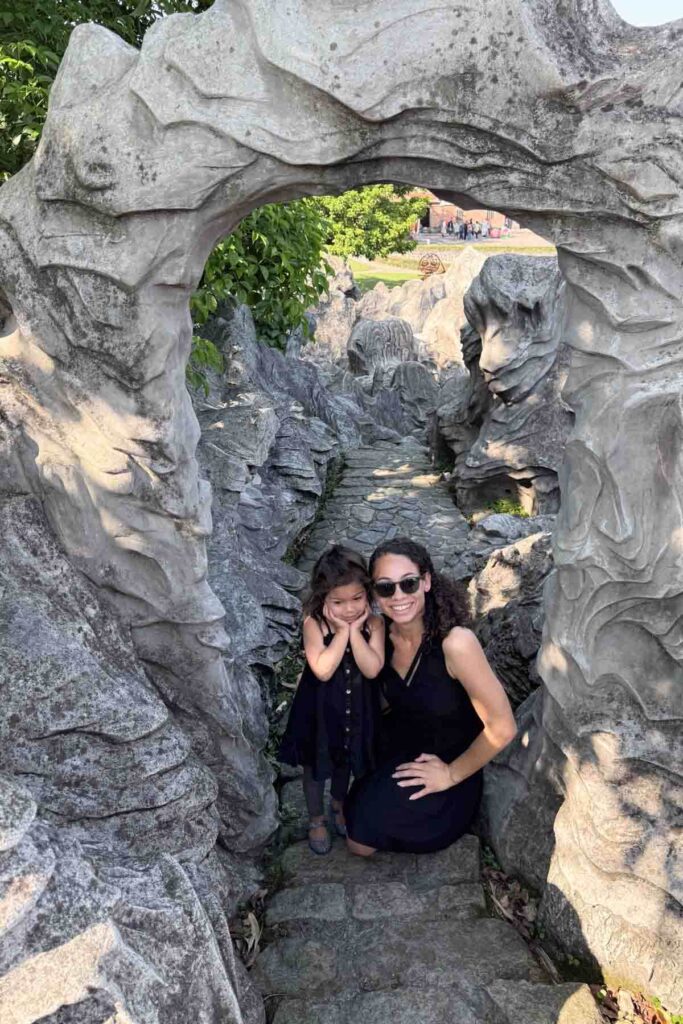 A woman and her toddler daughter pose under a carved stone archway in a rocky, landscaped path at the Lin An Tai Historical House Museum in Taipei. The stone appears weathered and sculptural, with lush greenery and traditional structures in the background.