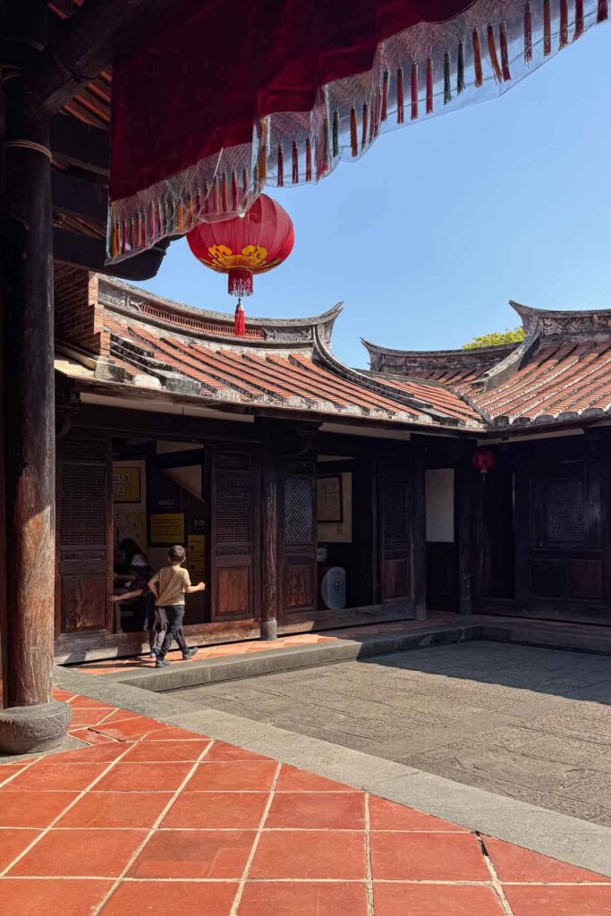 A bright red lantern hangs above a traditional courtyard with intricate wooden architecture and sweeping tiled roofs. A child runs toward an open doorway at the Lin An Tai Historical Mansion in Taipei, adding a sense of motion to the peaceful historic setting.