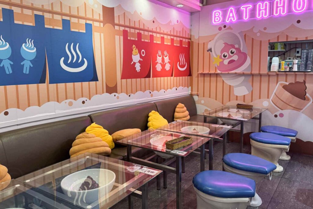 A whimsically decorated restaurant interior with cartoon poop decor, poop-shaped pillows, glass tables with toilet bowl dishes, and stools designed like toilet seats under a neon "BATH HOUSE" sign on a pastel-colored wall at the Modern Toilet Restaurant in Shilin, Taipei.