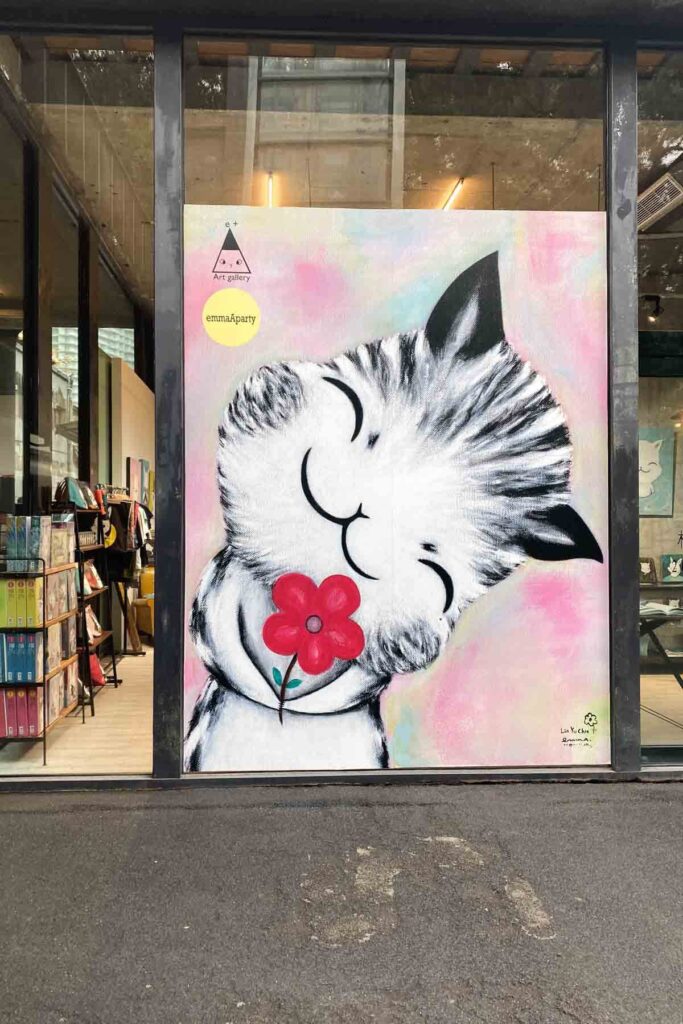 A colorful XL canvas painting of a black and white smiling cartoon cat holding a red flower in its paws, displayed on a wall of a shop at the Huashan 1914 Creative Park in Taipei.