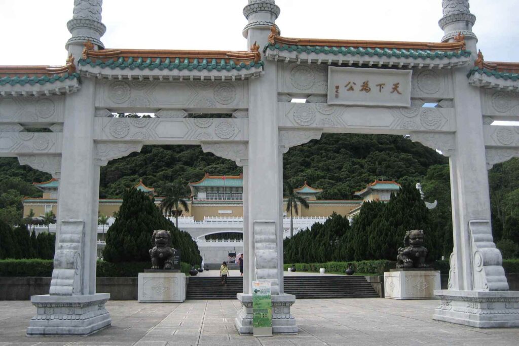 A grand white ceremonial gate with green and gold-tiled roofing frames the front view of the National Palace Museum in Taipei, set against a backdrop of dense greenery. Stone lion statues flank the entrance, enhancing the majestic scene.