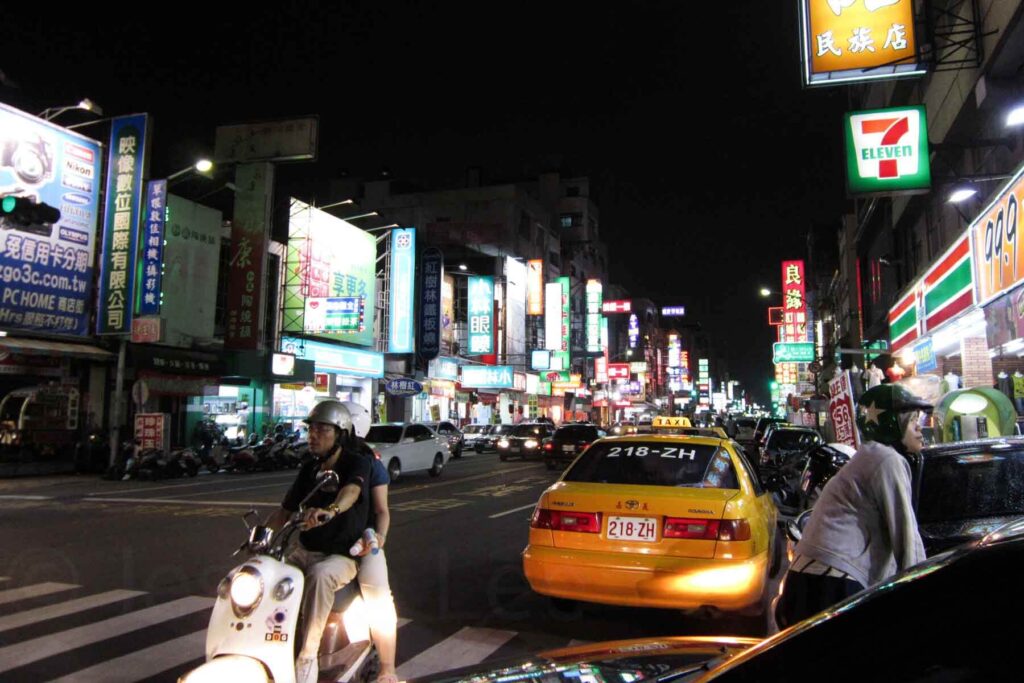 A bustling Taipei street at night is illuminated by neon signs and storefronts, with a yellow taxi, scooters, and pedestrians near a brightly lit 7-Eleven convenience store.