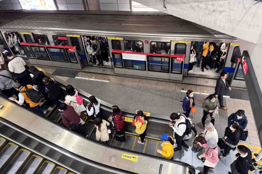 A bird's-eye view of a packed Taipei metro station shows commuters riding escalators and boarding a train. Many wear face masks, and platform signs indicate train routes and directions.