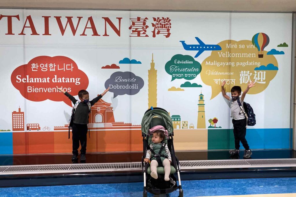 Three children, including one in a stroller, pose in front of a large multilingual welcome sign at Taoyuan International Airport in Taiwan, with “Taiwan” and “Welcome” written in various languages alongside playful travel-themed graphic illustrations.