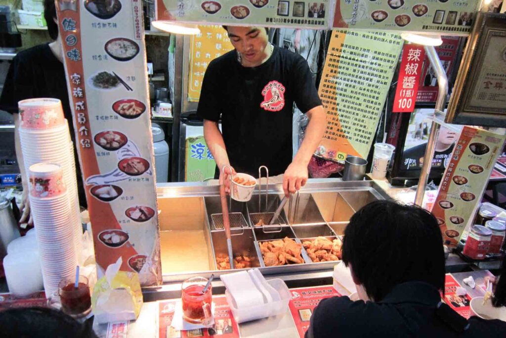 A food vendor prepares fried items behind a metal counter at a busy night market stall in Taiwan, with colorful signage in Chinese, menu images, and drinks placed in front of waiting customers.