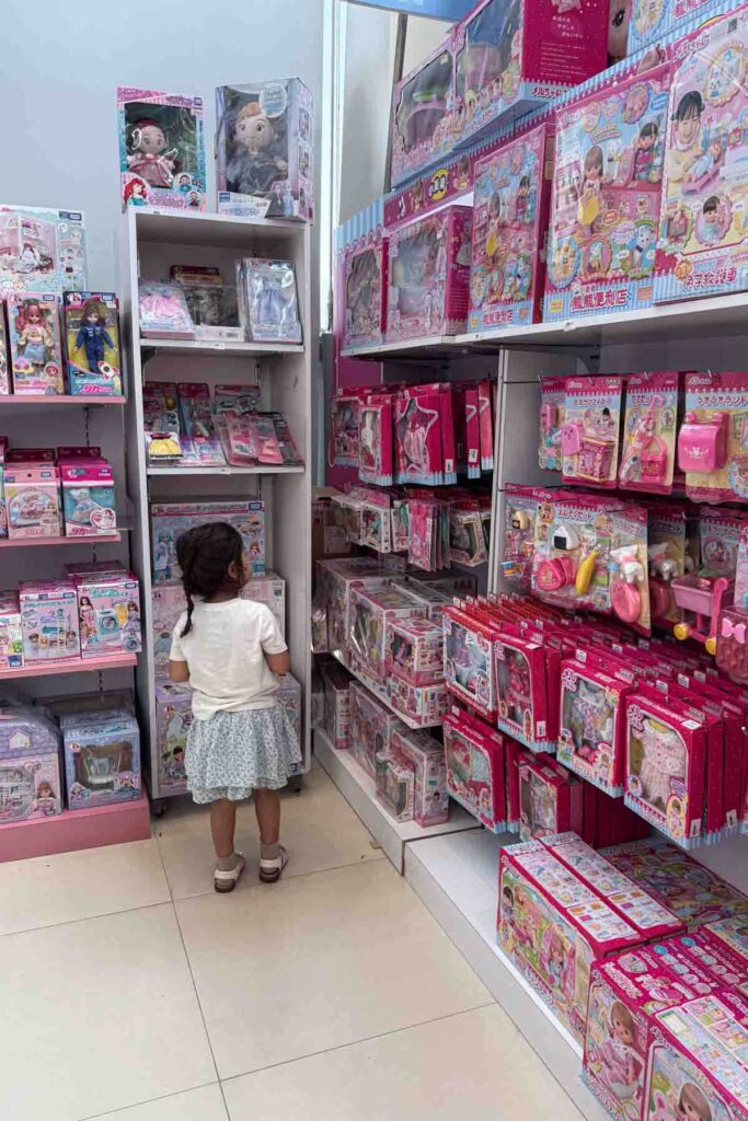 A little girl in a white top and floral skirt stands in front of shelves filled with pink toy sets and dolls in a brightly lit store. Boxes featuring Japanese writing and colorful packaging surround her in a SOGO store in Taipei.