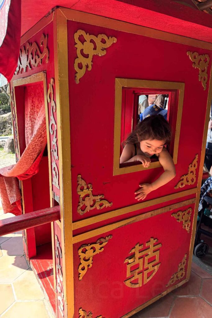 A young girl leans playfully out of the window of a vibrant red and gold traditional Chinese wedding sedan chair. Ornate golden patterns decorate the woodwork, and sunlight casts strong shadows across the tiled ground.