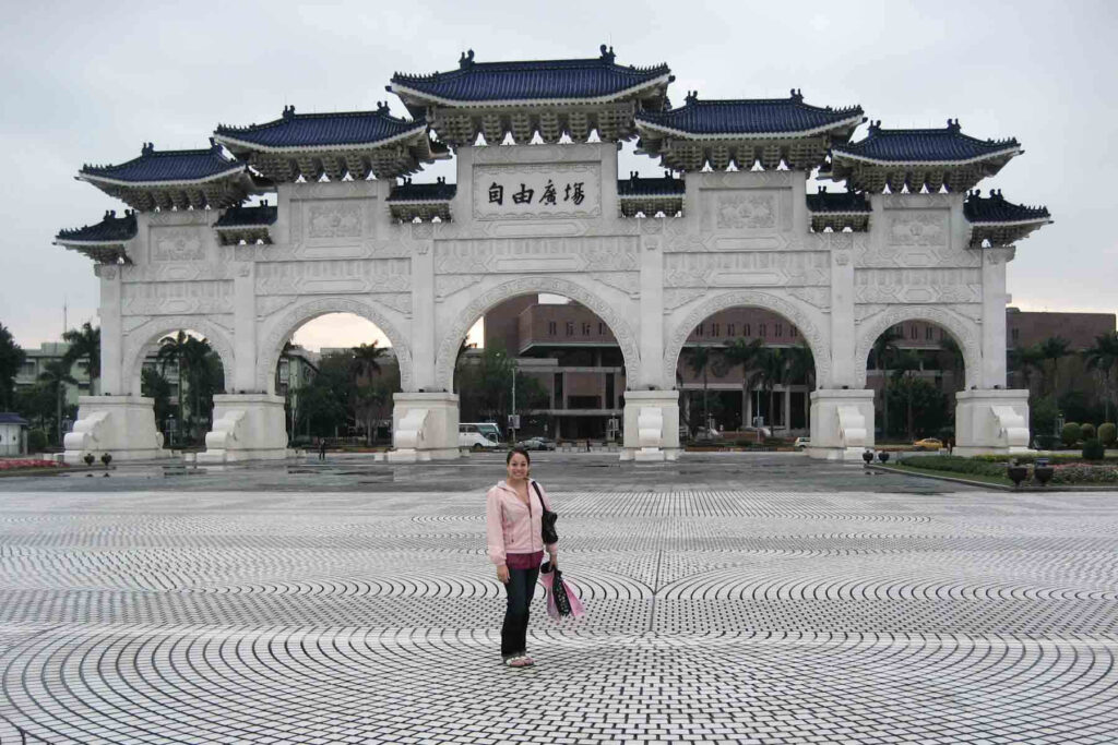 A smiling woman stands in front of Taipei’s iconic Liberty Square Arch, a striking white structure with blue-tiled roofs and intricate carvings. The patterned plaza beneath her feet adds visual depth to the historic landmark scene.
