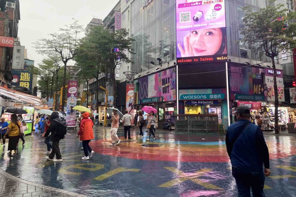 Shoppers carrying umbrellas walk through a rainbow, wet plaza in Ximending, Taipei. The Watsons storefront and large digital billboard overhead give the shopping district a lively, urban energy.