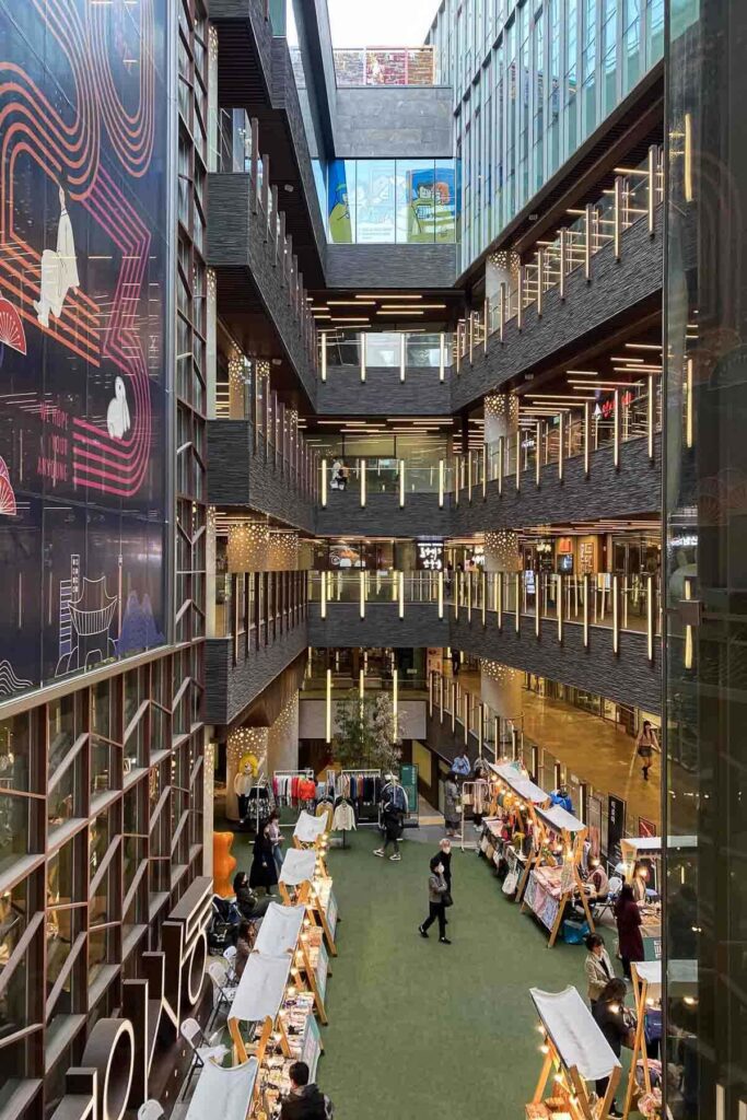 A multi-story indoor shopping mall, Anyoung in Insadong, with sleek glass walls with long bar lights, dark stone architecture, and rows of vendor booths on a green artificial turf floor, selling clothes, crafts, and food.