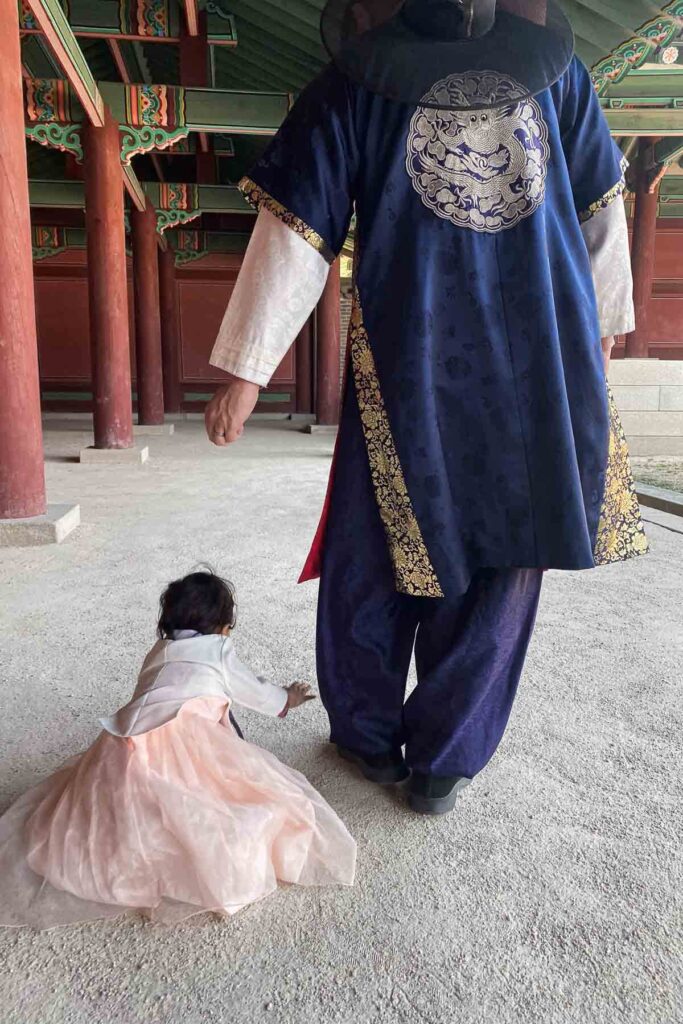 A toddler in a peach hanbok crawls on the ground beside an adult wearing a navy and gold traditional Korean hanbok inside a Changdeokgung palace corridor lined with red wooden columns.