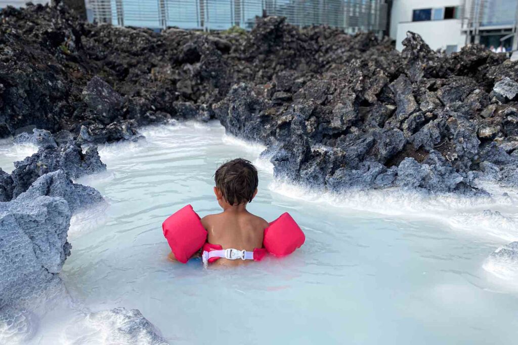 Toddler in pink puddle jumper crouches near white thickly covered silica rocks beside a bright blue geothermal pool at the Blue Lagoon.