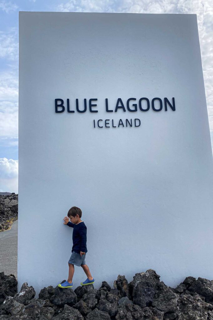 A young child stares up in awe at a giant puffin sculpture hanging above a baggage claim belt in the KEF Keflavík International Airport in Iceland, with travelers with suitcases and carts in the background.