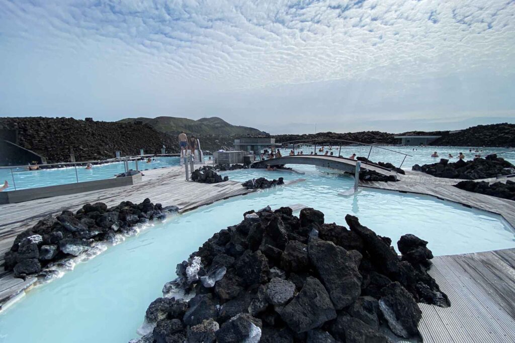 Sunny July day at the Blue Lagoon with people in the distance soaking in the mineral-rich water, framed by black lava rocks and steam rising in the background.