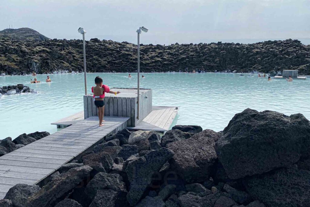 A toddler wearing floaties walks across a pale wooden deck near the edge of the Blue Lagoon with a large square wooden box at the end of the walkway flanked by tall lights on a pole, with calm blue water and volcanic rocks in the background.