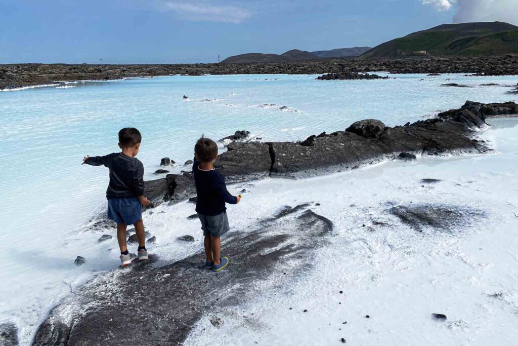 Two young brothers in long tees and shorts play along the lava rocks outside the Blue Lagoon, exploring the rugged terrain with bright blue geothermal water behind them.