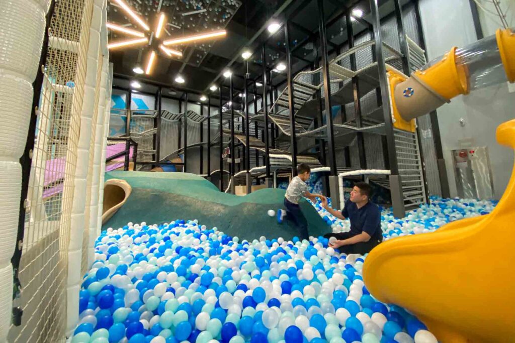 A spacious, modern indoor play area, Champion 1250, in IPark Mall in Seoul, features a massive ball pit filled with blue and white balls, a green climbing island, a yellow slide, netted climbing structures, and a father helping his child climb down into the balls.
