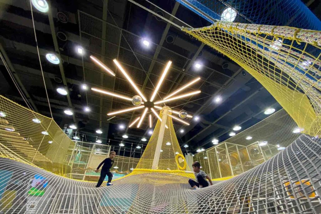 Two children play on a large yellow and gray net structure under dramatic ceiling lights shaped like a starburst, inside Champion 1250, a modern kids’ indoor playground in Seoul, located in IPark Mall.