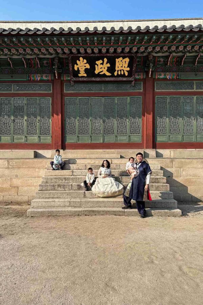 A young family of 5 dressed in traditional Korean hanbok sits on the stone steps of the historic Changgyeonggung palace building with green latticed doors and a golden sign above them.