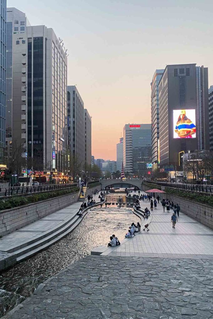 A wide view of Cheonggyecheon Stream in downtown Seoul, Korea at sunset, with people strolling or sitting along the waterway between rows of modern skyscrapers and bright digital billboards.