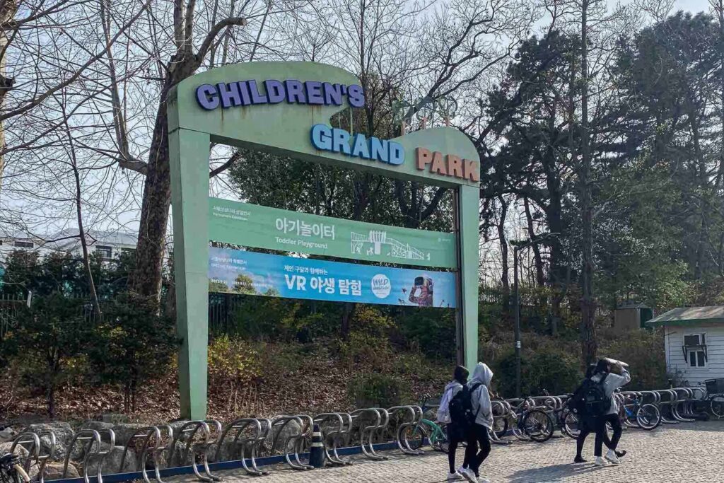 The entrance to Children’s Grand Park in Seoul marked by a pastel green archway with colorful letters and signs in both Korean and English, with a bike rack and groups of people walking nearby.