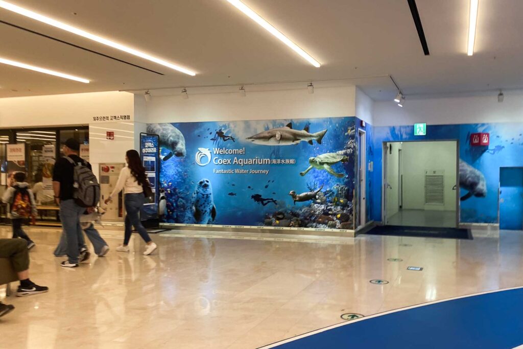 A family walks past a large blue wall mural at the Coex Aquarium entrance in Seoul, inside a mall, featuring marine animals like sharks, turtles, and seals with the slogan “Fantastic Water Journey.”