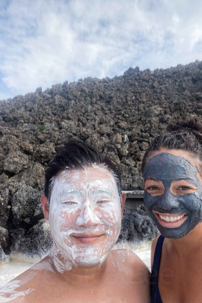 Smiling couple with facial masks at the Blue Lagoon in Iceland, one covered in a white silica mask and the other in a dark mud mask, standing in front of a lava rock wall with the spa waters just below.