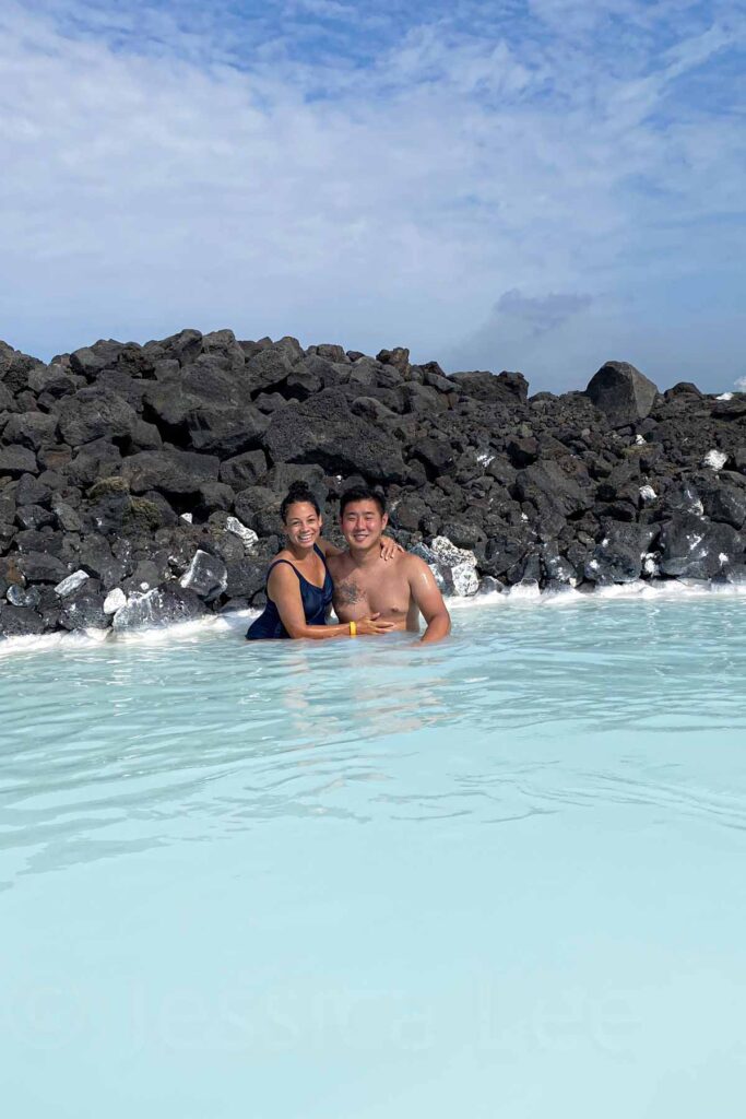 A smiling couple waist-deep in the milky blue aqua waters of the Blue Lagoon in Iceland, with dark volcanic rocks lining the shore and a bright blue sky above.