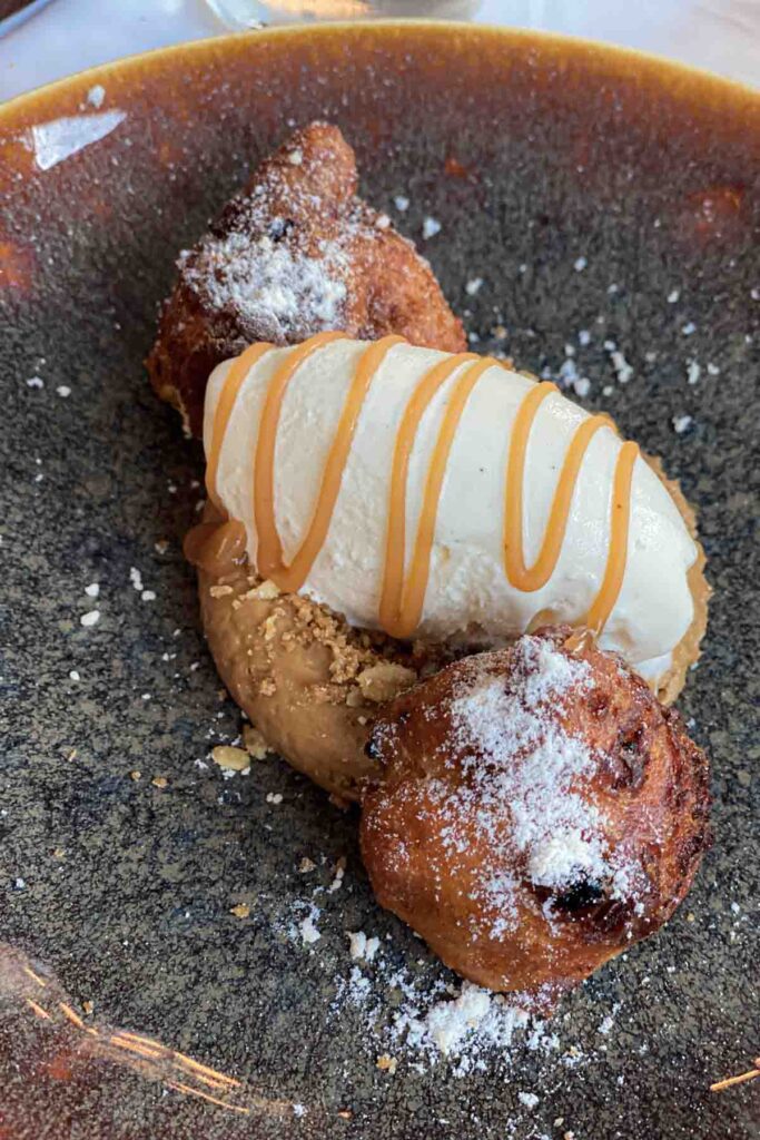 Plated dessert featuring a scoop of vanilla ice cream drizzled with caramel, served with small fried pastries dusted in powdered sugar and a nutty crumble base served at the Lava Restaurant.