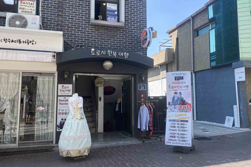 A hanbok rental store, Dorothy Hanbok in Seoul, with a mannequin in a pale blue and gold hanbok stands next to a sign displaying rental pricing and photos of visitors in traditional attire.