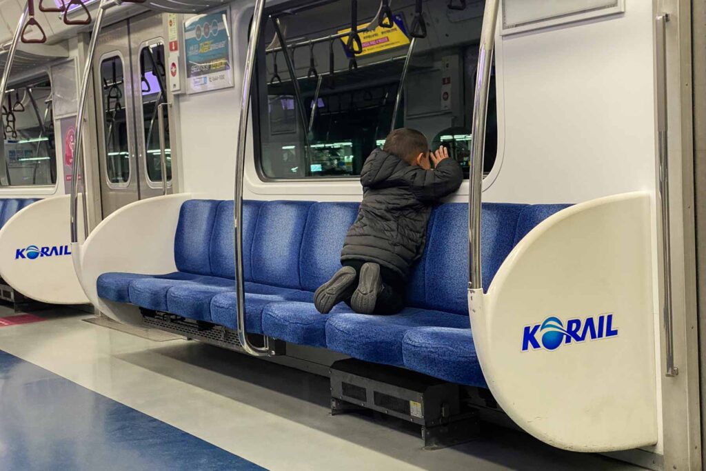 A young boy in a black puffer jacket kneels on a blue subway seat labeled “KORAIL” while peering out the window of an empty train car in Seoul, South Korea.