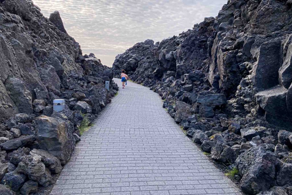 A young child walks ahead on a stone path cutting through rugged lava rock formations, creating a dramatic pathway leaving the Blue Lagoon in Iceland.