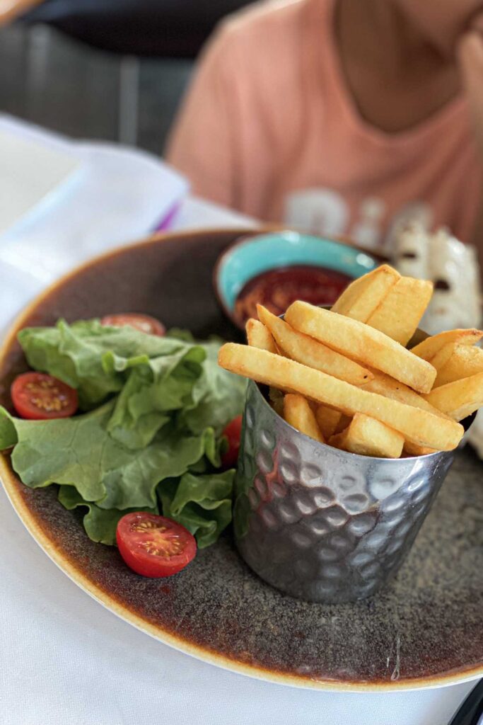 Kid’s meal plate with thick-cut fries in a metal cup, fresh lettuce, cherry tomatoes, ketchup, and a partially visible wrap, with a child in a light orange tee sitting in the background.