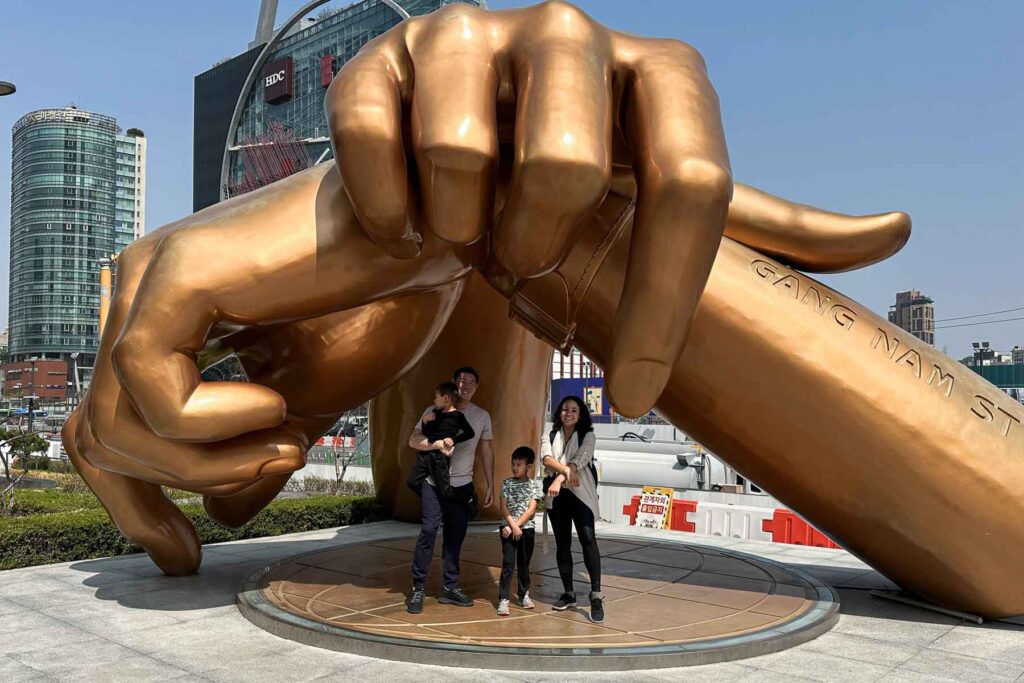 A family poses in front of COEX Mall beneath a giant bronze sculpture of crossed hands performing the Gangnam Style dance gesture, set against a modern cityscape in Seoul.