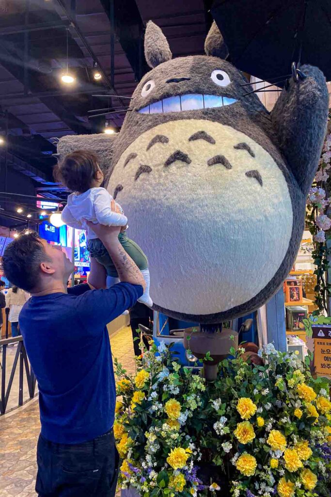 A father lifts his toddler toward a giant plush statue of Totoro holding an umbrella, surrounded by yellow flowers and indoor lighting in a themed shopping space at IPark Mall in Seoul.